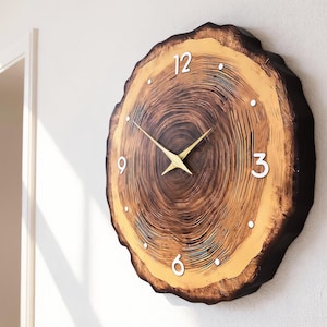 Puede incluir: Un reloj de pared redondo diseñado para parecerse a una sección transversal de un tronco de árbol. La esfera del reloj presenta un patrón de vetas de madera natural con números blancos en las posiciones de las 12, 3, 6 y 9 en punto. Las manecillas del reloj son doradas.