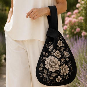 May include: A black fabric handbag with a teardrop shape and a single wrist strap. The bag features a detailed, light-colored floral print of large peonies and leaves covering the front surface, creating a high-contrast botanical design.