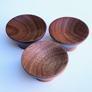 May include: Three small, round wooden bowls with a dark brown finish. The bowls are made of a dark wood with a natural grain pattern. They are all the same size and shape, and they are arranged in a triangular pattern.