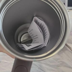 May include: A stainless steel travel mug with a gray handle. Inside the mug is a folded white paper with black text. The mug is empty and ready to be filled with a beverage.