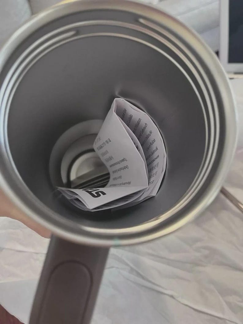 May include: A stainless steel travel mug with a gray handle. Inside the mug is a folded white paper with black text. The mug is empty and ready to be filled with a beverage.