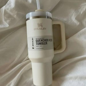Puede incluir: Vaso Stanley Quencher H2.0 color crema con asa y pajita. El vaso tiene el logotipo de Stanley y el texto "THE FLOWSTATE QUENCHER H2.0 TUMBLER" y "40 OZ | 1.18L". El vaso est&aacute; sobre una superficie blanca.