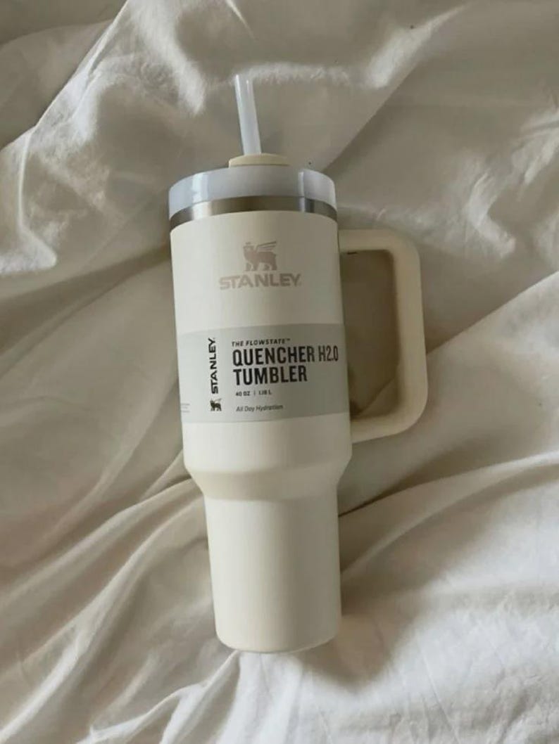 Puede incluir: Vaso Stanley Quencher H2.0 color crema con asa y pajita. El vaso tiene el logotipo de Stanley y el texto "THE FLOWSTATE QUENCHER H2.0 TUMBLER" y "40 OZ | 1.18L". El vaso est&aacute; sobre una superficie blanca.