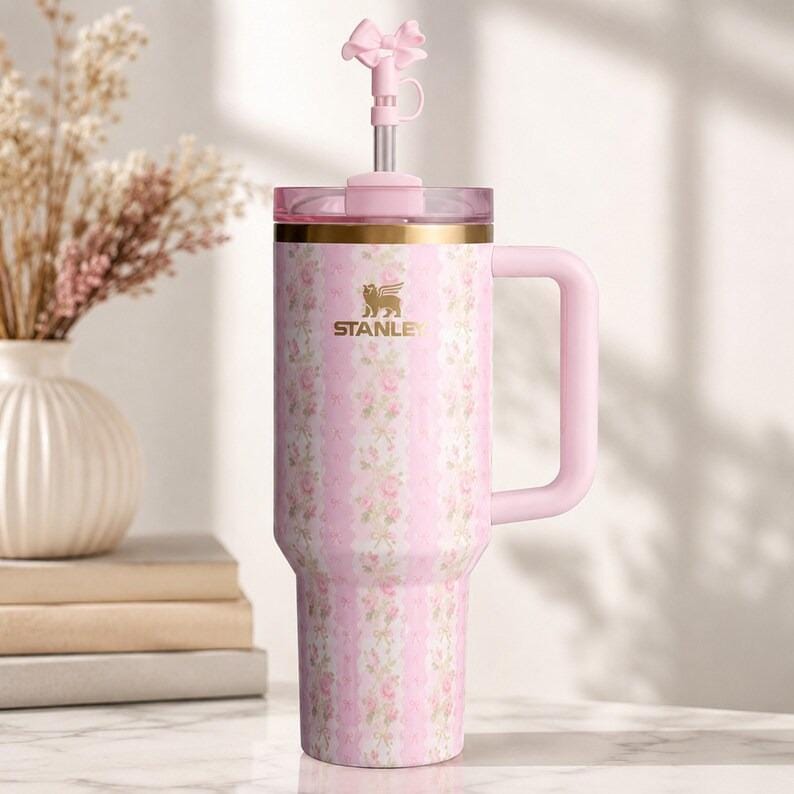 Peut inclure: Gobelet Stanley rose et blanc &agrave; motifs floraux, avec une poign&eacute;e et un bouchon de paille en forme de n&oelig;ud. Le gobelet a un bord dor&eacute; et le logo Stanley. Le motif floral est &agrave; rayures verticales avec des roses.