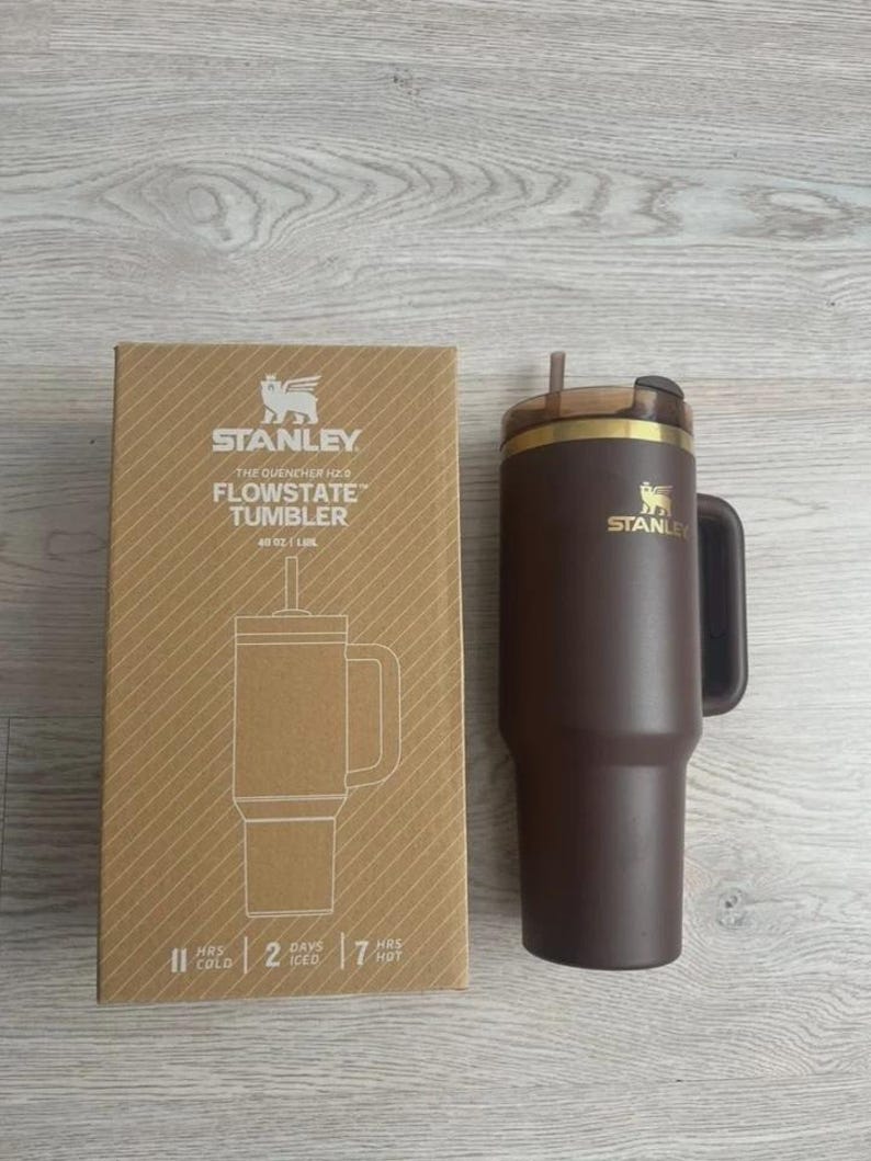 May include: A brown Stanley Flowstate tumbler with a handle and straw, next to its cardboard box. The box features the Stanley logo and product details, including "40 oz". The tumbler is designed to keep drinks cold for 2 days and hot for 7 hours.