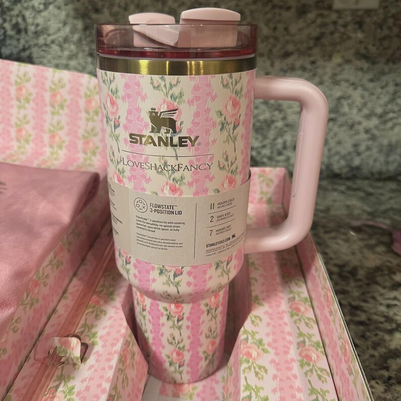May include: A pink and floral Stanley Loveshackfancy Flowstate tumbler with a handle. The tumbler has a pink lid and a floral pattern with the Stanley logo. The tumbler is in a matching floral patterned box.
