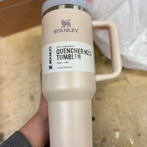 Puede incluir: Un vaso Stanley Quencher H2.0 rosa claro con asa y tapa azul claro. El vaso tiene el logotipo de Stanley y el texto "THE FLOWSTATE QUENCHER H2.0 TUMBLER" impreso en el lateral. Contiene 1,18 litros.