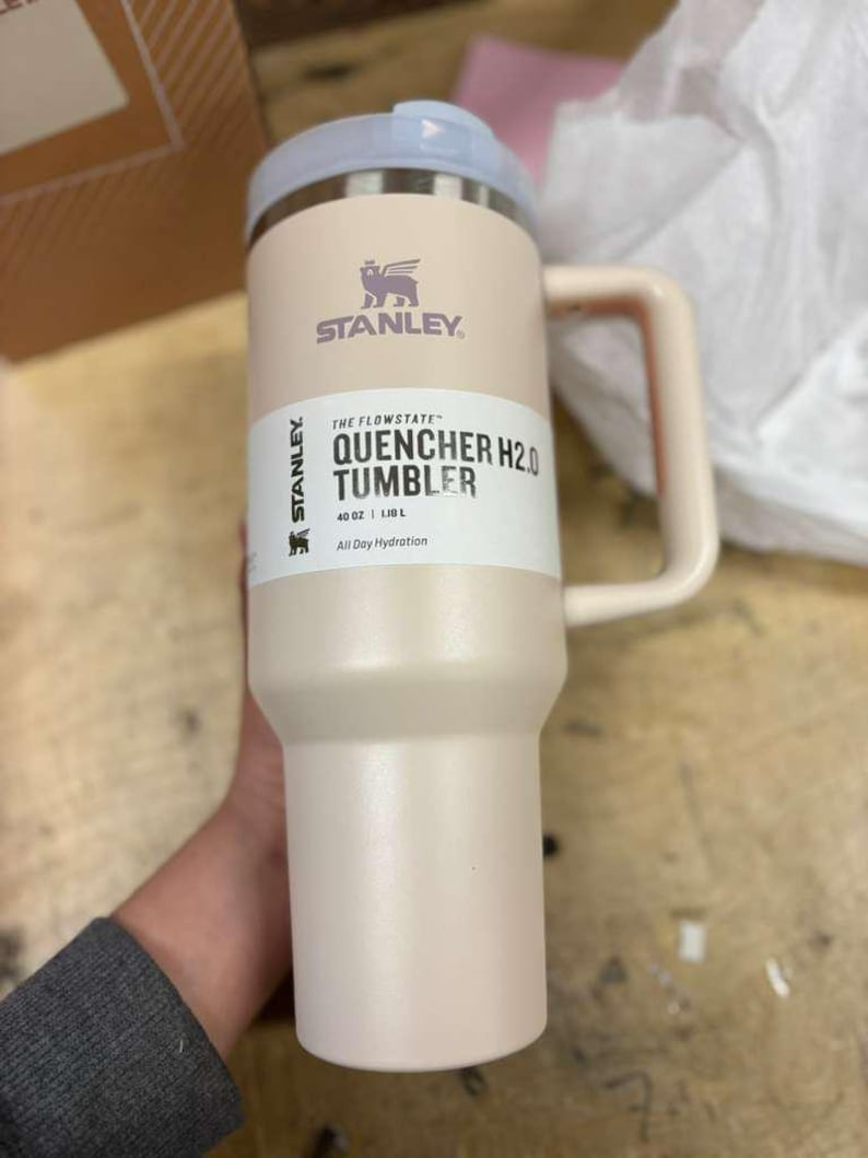 Puede incluir: Un vaso Stanley Quencher H2.0 rosa claro con asa y tapa azul claro. El vaso tiene el logotipo de Stanley y el texto "THE FLOWSTATE QUENCHER H2.0 TUMBLER" impreso en el lateral. Contiene 1,18 litros.