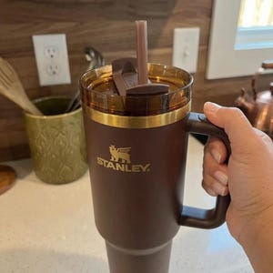 May include: A brown Stanley tumbler with a handle, gold trim, and a straw. The Stanley logo is visible on the front. The tumbler is held by a person's hand.
