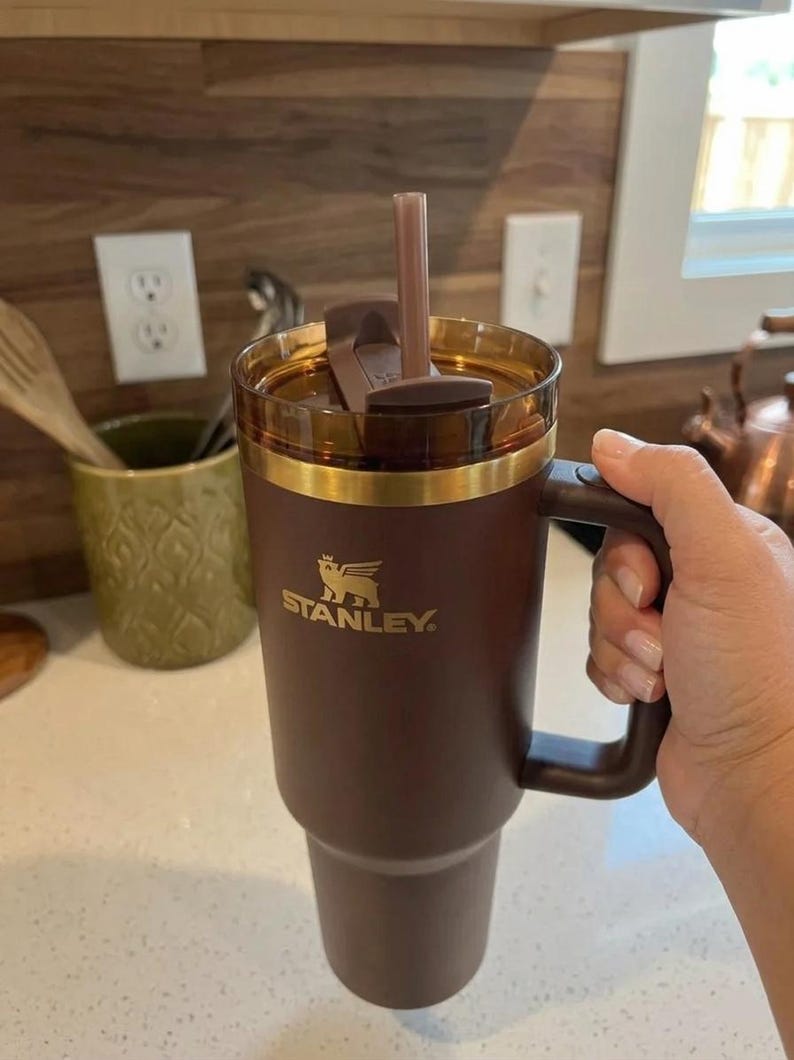May include: A brown Stanley tumbler with a handle, gold trim, and a straw. The Stanley logo is visible on the front. The tumbler is held by a person's hand.
