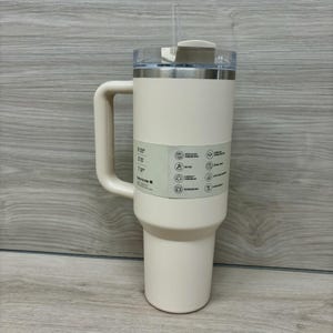 Puede incluir: Un vaso t&eacute;rmico de color crema con asa y tapa transparente. El vaso tiene un borde de acero inoxidable y una pajita. Una etiqueta en el vaso muestra iconos y texto. El vaso est&aacute; dise&ntilde;ado para mantener las bebidas fr&iacute;as o calientes.