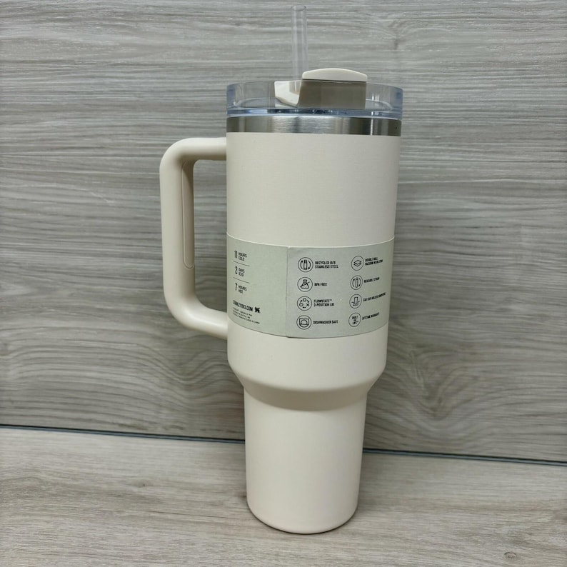 Puede incluir: Un vaso t&eacute;rmico de color crema con asa y tapa transparente. El vaso tiene un borde de acero inoxidable y una pajita. Una etiqueta en el vaso muestra iconos y texto. El vaso est&aacute; dise&ntilde;ado para mantener las bebidas fr&iacute;as o calientes.