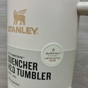 Puede incluir: Un vaso blanco Stanley Quencher H2.0 con asa. El vaso tiene el logotipo de Stanley y el texto "THE FLOWSTATE QUENCHER H2.0 TUMBLER". Una pegatina redonda dice "Hearth & Hand with Magnolia LIMITED EDITION COLOR".