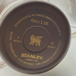 May include: A brown Stanley brand water bottle base with gold text. The text reads "Designed in Seattle WA", "40oz / 1.18L", "Dishwasher Safe", and "Do Not Microwave". A gold logo of a winged creature is in the center.