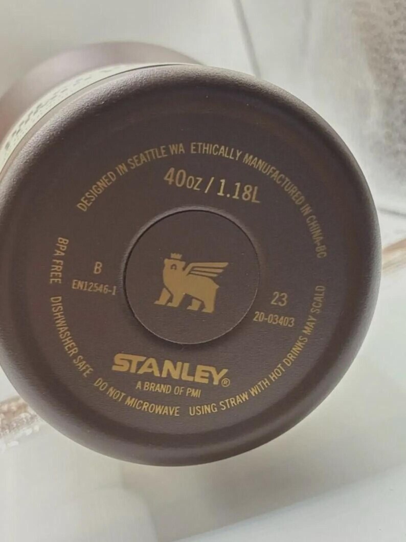 May include: A brown Stanley brand water bottle base with gold text. The text reads "Designed in Seattle WA", "40oz / 1.18L", "Dishwasher Safe", and "Do Not Microwave". A gold logo of a winged creature is in the center.