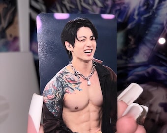 May include: A rectangular photo card featuring a smiling person with dark hair, wearing a silver chain necklace and an open dark jacket. The person has visible tattoos on their right arm and a muscular torso, captured in a concert setting.