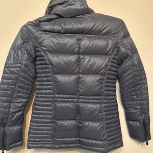 Puede incluir: Una chaqueta acolchada gris con capucha. La chaqueta tiene un dise&ntilde;o acolchado en la espalda y las mangas, con paneles acanalados verticales en los laterales. Cuenta con cierre de cremallera y cuello alto.