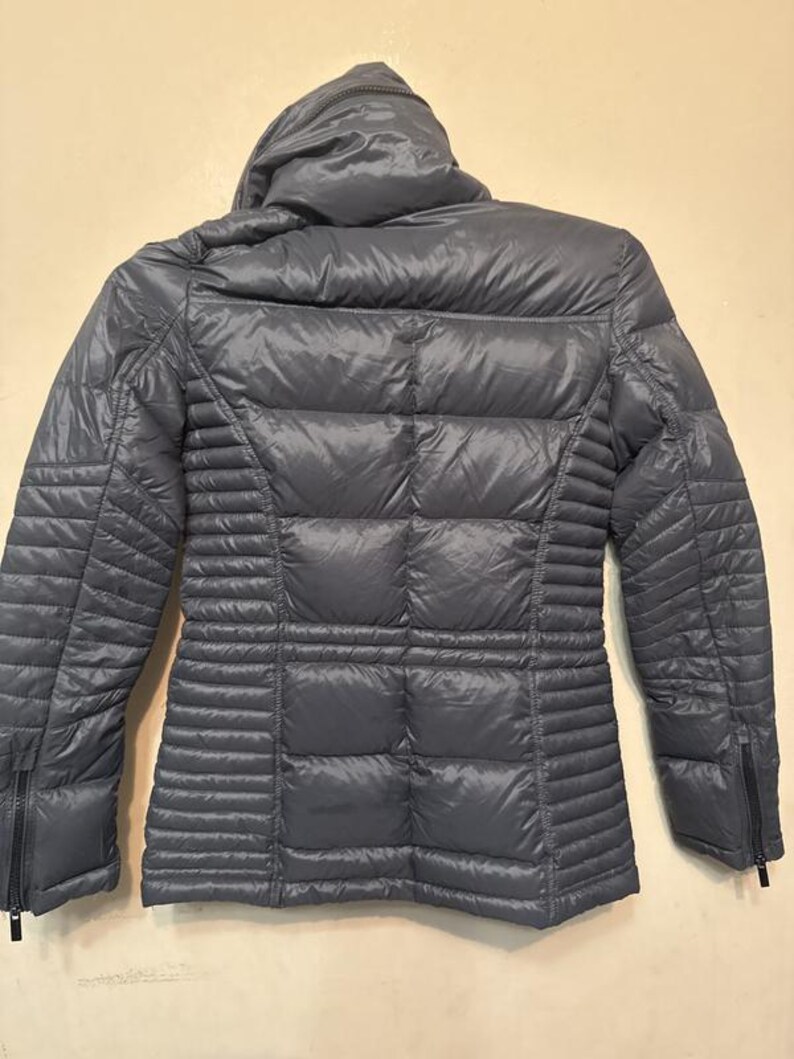 Puede incluir: Una chaqueta acolchada gris con capucha. La chaqueta tiene un dise&ntilde;o acolchado en la espalda y las mangas, con paneles acanalados verticales en los laterales. Cuenta con cierre de cremallera y cuello alto.