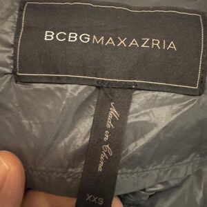 Puede incluir: Primer plano de una etiqueta negra de una chaqueta BCBGMAXAZRIA. La etiqueta tiene el nombre de la marca en letras may&uacute;sculas blancas. Una etiqueta m&aacute;s peque&ntilde;a debajo dice "Made in China" y "XXS".