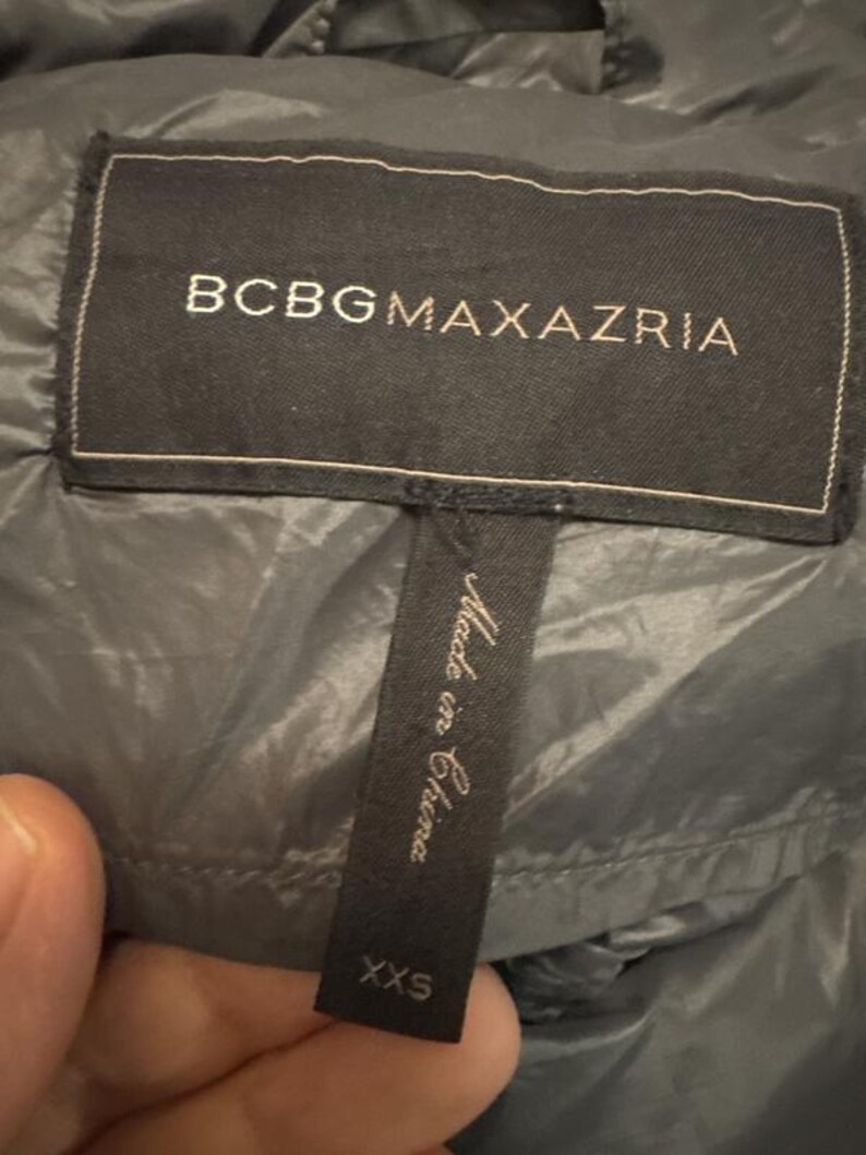Puede incluir: Primer plano de una etiqueta negra de una chaqueta BCBGMAXAZRIA. La etiqueta tiene el nombre de la marca en letras may&uacute;sculas blancas. Una etiqueta m&aacute;s peque&ntilde;a debajo dice "Made in China" y "XXS".