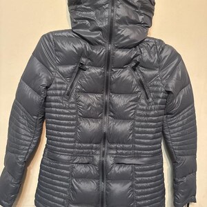 Puede incluir: Una chaqueta acolchada gris con cuello alto y cremallera completa. La chaqueta tiene dos bolsillos con cremallera y un dise&ntilde;o acolchado. Las mangas son largas y la chaqueta parece estar hecha de un material ligero y resistente al agua.