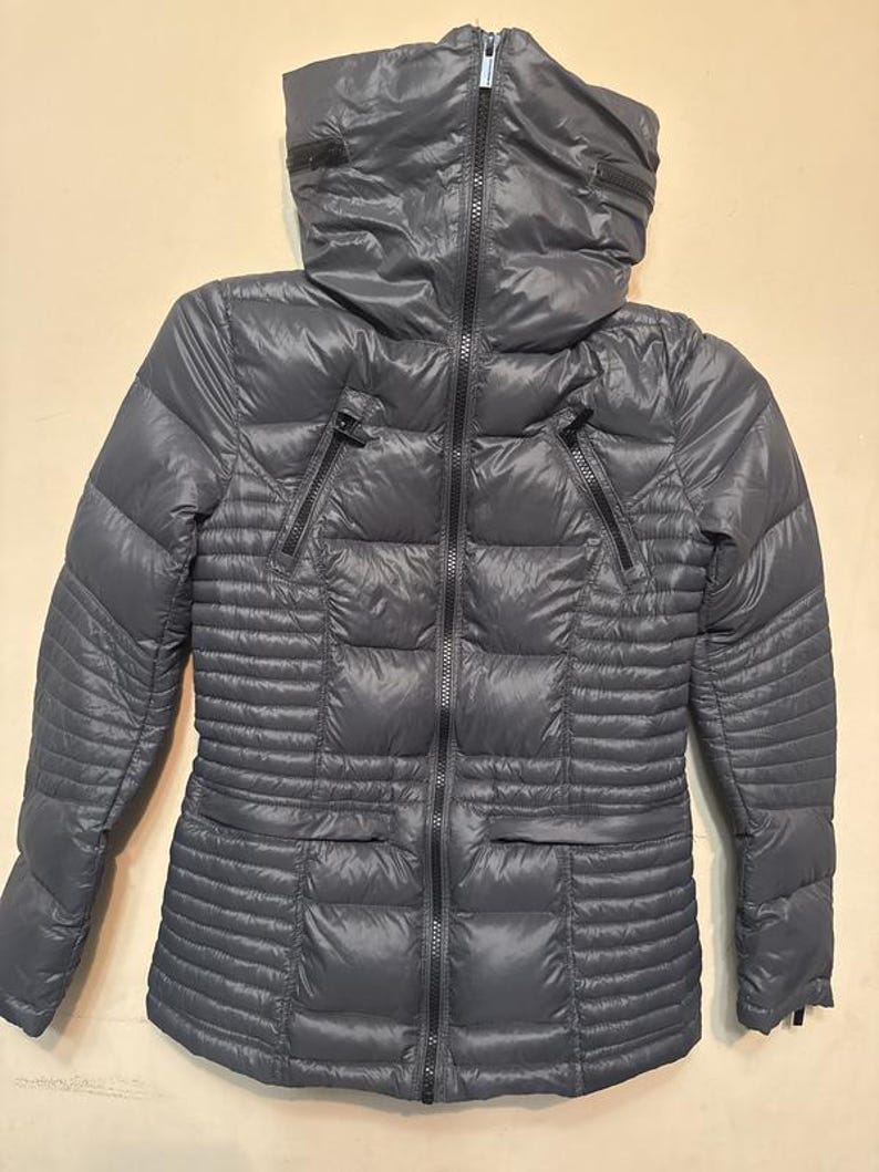 Puede incluir: Una chaqueta acolchada gris con cuello alto y cremallera completa. La chaqueta tiene dos bolsillos con cremallera y un dise&ntilde;o acolchado. Las mangas son largas y la chaqueta parece estar hecha de un material ligero y resistente al agua.