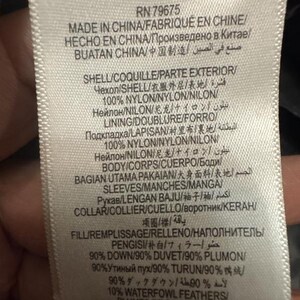 Puede incluir: Una etiqueta de ropa blanca con texto negro, que detalla la composici&oacute;n y el origen de la prenda. La etiqueta incluye informaci&oacute;n en varios idiomas, como ingl&eacute;s, franc&eacute;s y chino, e indica que los materiales son 90% plum&oacute;n y 10% plumas de aves acu&aacute;ticas.