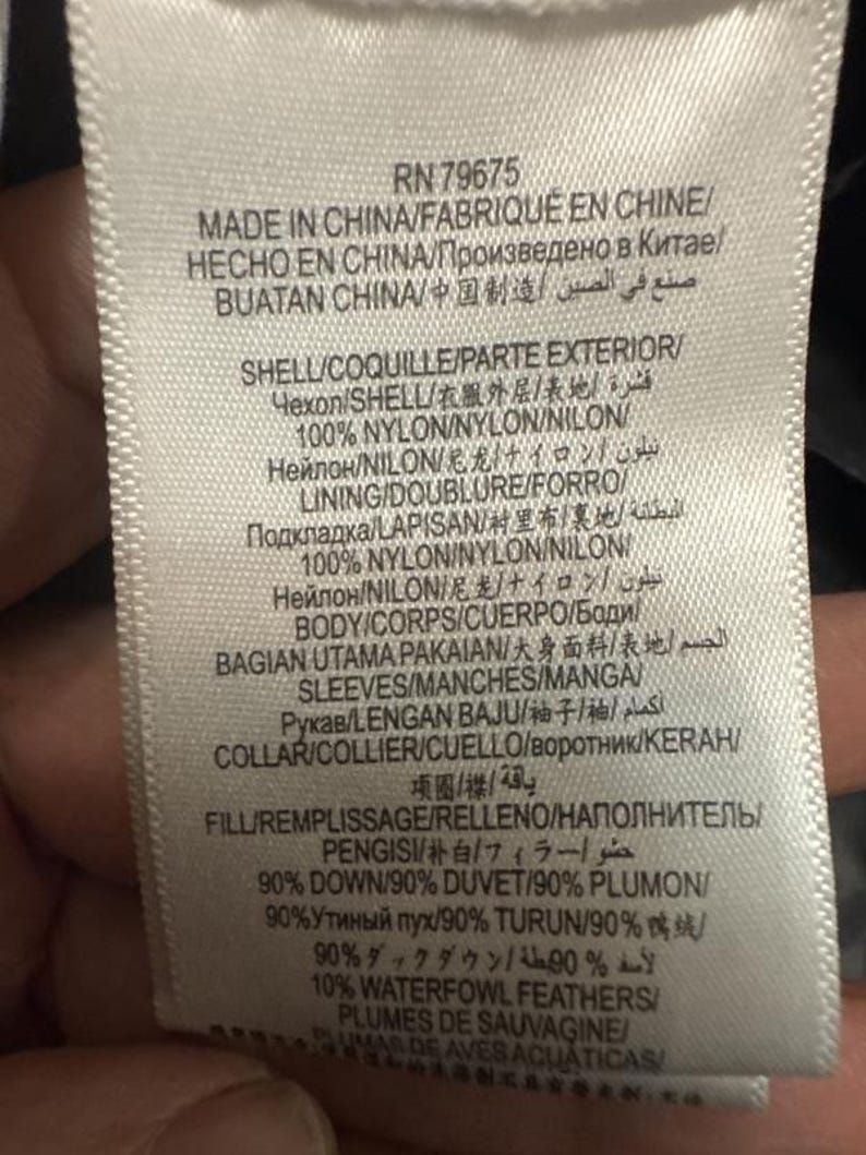Puede incluir: Una etiqueta de ropa blanca con texto negro, que detalla la composici&oacute;n y el origen de la prenda. La etiqueta incluye informaci&oacute;n en varios idiomas, como ingl&eacute;s, franc&eacute;s y chino, e indica que los materiales son 90% plum&oacute;n y 10% plumas de aves acu&aacute;ticas.