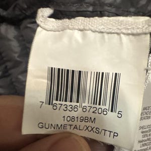 Puede incluir: Primer plano de una etiqueta de ropa con un c&oacute;digo de barras y el texto "GUNMETAL/XXS/TTP". La etiqueta es blanca con texto negro. El fondo es una tela acolchada gris oscuro.