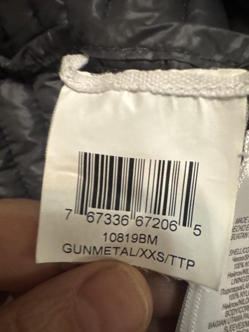 Puede incluir: Primer plano de una etiqueta de ropa con un c&oacute;digo de barras y el texto "GUNMETAL/XXS/TTP". La etiqueta es blanca con texto negro. El fondo es una tela acolchada gris oscuro.