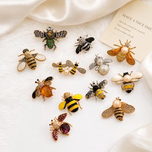 May include: A collection of decorative bee brooches in various designs. The brooches feature a variety of colors, including gold, black, yellow, and red, with some adorned with pearls and crystals. The image also includes a card with the text "HAVE A NICE DAY."