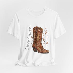Könnte beinhalten: Weißes T-Shirt mit einer detaillierten Illustration eines braunen und goldenen Cowboy-Stiefels. Der Stiefel hat ein Blitzdesign und ist von Sternen und Blitzen umgeben. Der Text auf dem Shirt lautet "Disco Desert."