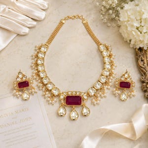 Puede incluir: Un conjunto de collar y pendientes en tono dorado con piedras rectangulares de color magenta intenso, cristales transparentes en forma de lágrima y pequeñas cuentas blancas tipo perla. El collar tiene una cadena de varios hilos y un colgante central con tres cristales colgantes.