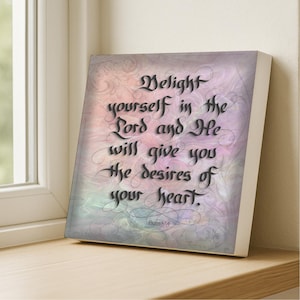 May include: A square wall art piece featuring the black calligraphic text "Delight yourself in the Lord and He will give you the desires of your heart. Psalm 37:4" set against a soft, swirling background of blended pastel pink, purple, and blue hues.