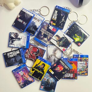 Puede incluir: Una colección de llaveros rectangulares en miniatura con varias portadas de videojuegos, incluyendo títulos como Resident Evil 4, The Last of Us, Call of Duty, The Witcher, Cyberpunk, Minecraft y Grand Theft Auto, cada uno con una anilla metálica.