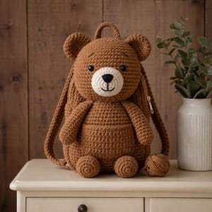 May include: A brown crocheted bear backpack with a white muzzle and black eyes. The backpack has a zipper closure, adjustable straps, and a small front pocket. A ball of brown yarn sits next to the backpack. The bear has a friendly expression.