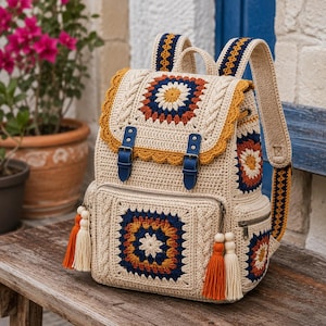 Op de afbeelding: Crèmekleurige gehaakte rugzak met een vierkant bloemmotief in marineblauw, oranje en geel. De rugzak heeft een klep met blauwe gespen, een voorvak en oranje en crèmekleurige kwastjes. De bandjes en de afwerking hebben een decoratief patroon.