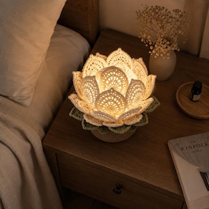 May include: A handcrafted lotus flower lamp with an intricate crochet design. The cream and green lamp emits a warm light, creating a cosy atmosphere. It sits on a wooden bedside table. The lamp is approximately 25 cm in diameter.