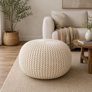 May include: A cream-coloured knitted pouf, with a visible textured knit pattern, sits on a neutral-toned rug. The background includes a sofa, a coffee table, and a woven basket with a plant, creating a cosy living room setting. The pouf is the focal point.
