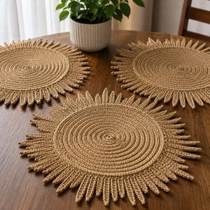 Puede incluir: Tres salvamanteles redondos de ganchillo beige con diseño de sol dispuestos sobre una mesa de madera. Cada salvamanteles presenta un centro en espiral y flecos radiantes. Una planta en maceta se encuentra en el fondo.