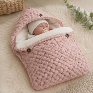May include: A sleeping baby wearing a white beanie and pacifier is nestled in a pink, woven-textured baby sleeping bag. The sleeping bag has a hood and is secured with two brown buttons. The interior is white.