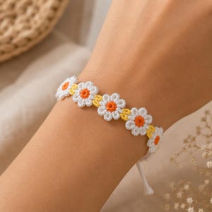 May include: A daisy chain bracelet featuring white crochet flowers with orange centers and yellow accents. The bracelet is tied with a white string. The bracelet is a charming accessory.