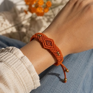 May include: An orange macrame bracelet with a diamond pattern and small gold beads. The bracelet is tied with an adjustable orange cord. The bracelet is on a person's wrist, with a cream-colored sweater and blue jeans in the background.