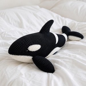 Peut inclure: Une peluche d'orque crochetée, noire et blanche. Le jouet en peluche a un aileron dorsal et des nageoires caudales. L'orque a des marques blanches autour des yeux et sur le ventre. Le jouet est posé sur un lit blanc.