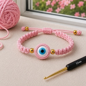 May include: Pink macrame bracelet featuring a blue eye charm with gold beads. The bracelet is adjustable and sits next to a ball of pink yarn and a 2.5 mm crochet hook. The background includes a window with pink flowers.