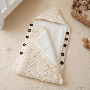 Puede incluir: Saco para bebé de color crema con capucha y botones marrones oscuros. La capa exterior de punto presenta un diseño de trenzas, mientras que el interior está forrado con forro polar blanco y suave. Una manta de envoltura cálida y acogedora.