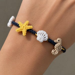 Op de afbeelding: Gehaakte armband met een zeester, schelp en zeepaardje. De armband heeft een marineblauwe koord met gele accenten. De zeester is geel, de schelp is wit en het zeepaardje is beige. Nautisch thema.