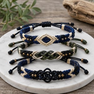 May include: Three macrame bracelets in navy blue, olive green, and black, each with intricate woven patterns and adjustable cords. The bracelets feature geometric designs and small gold-colored beads, displayed on a white marble surface.