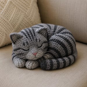 May include: A crocheted cat figurine in a curled-up sleeping position. The cat is gray with darker gray stripes and a pink nose. The handmade cat is resting on a beige surface, with a beige pillow in the background.
