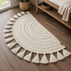May include: A half-circle, off-white crochet rug with a detailed circular pattern. The rug is trimmed with multiple tassels along the curved edge. The rug is placed on a wooden floor.